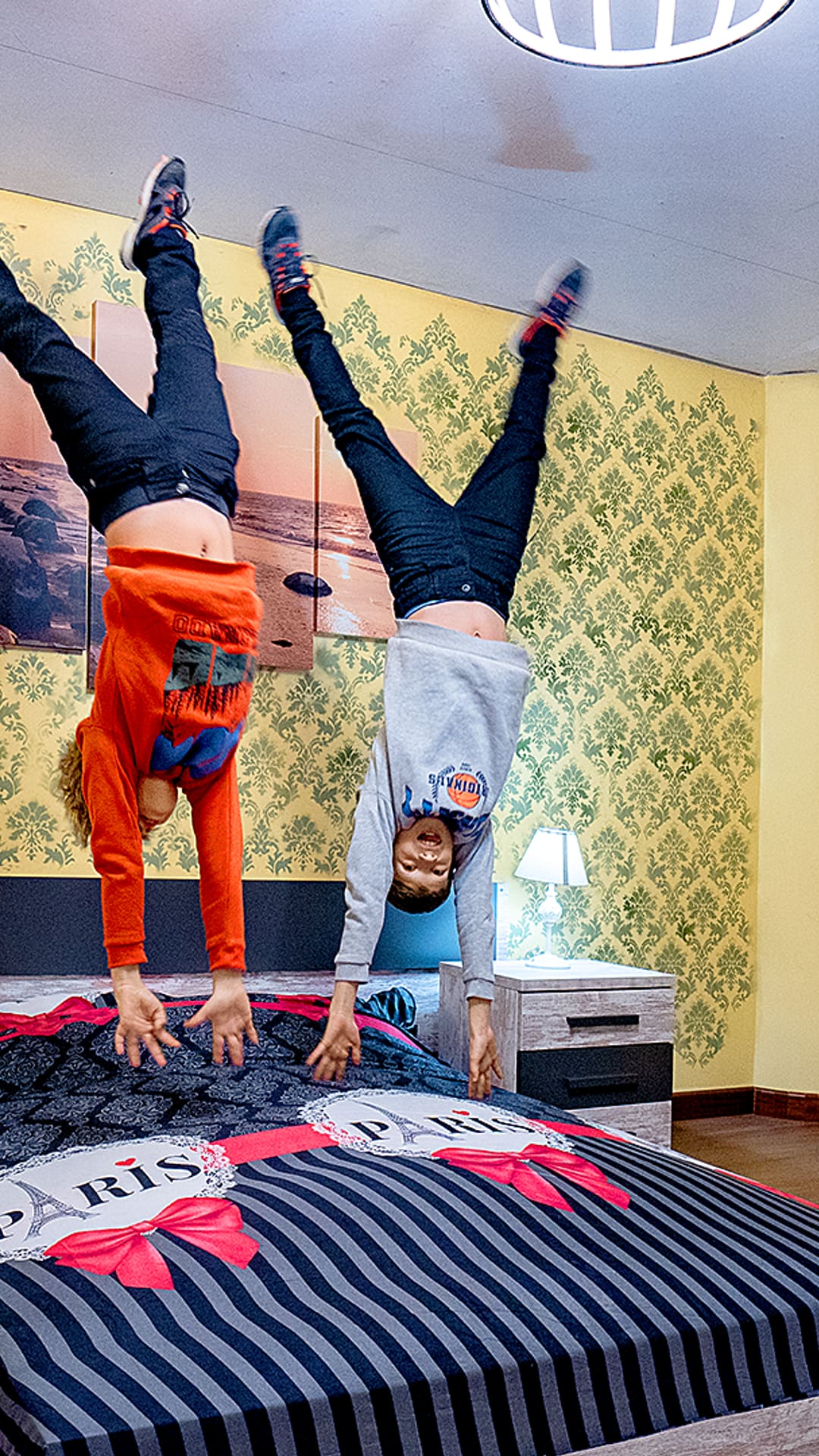 Two children doing handstands upside down on a striped Paris-themed bed in an inverted bedroom at Upside Down House Barcelona
