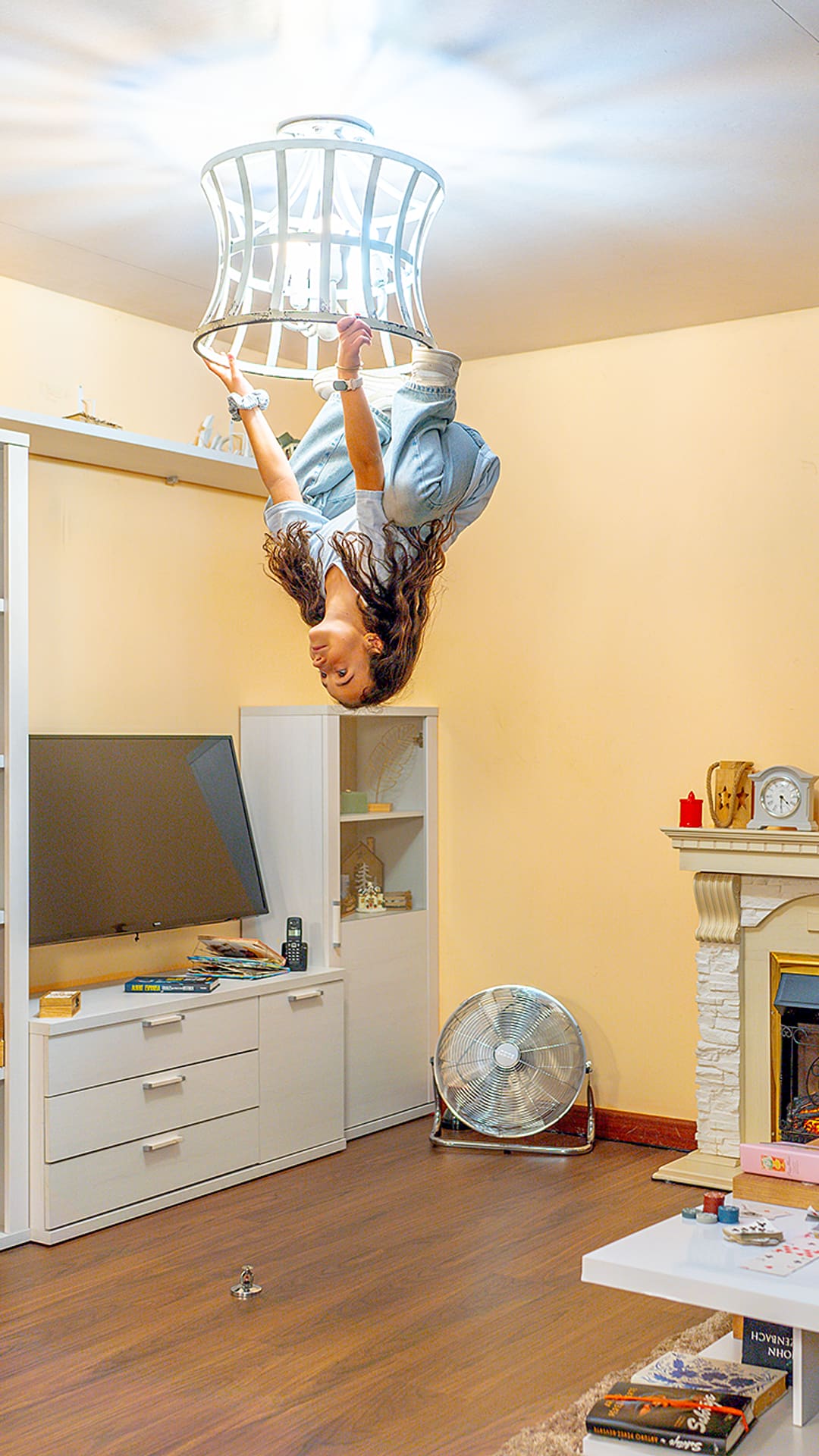 Woman hanging upside down from a ceiling light fixture above an inverted living room with a fireplace and TV at Upside Down House Barcelona